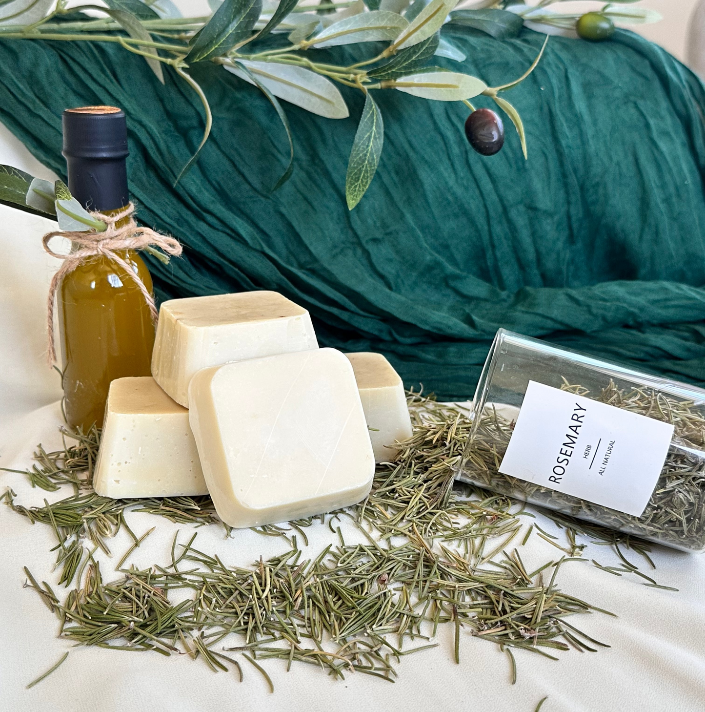 Herbal soap bars and a bottle of oil on a white surface with rosemary leaves.