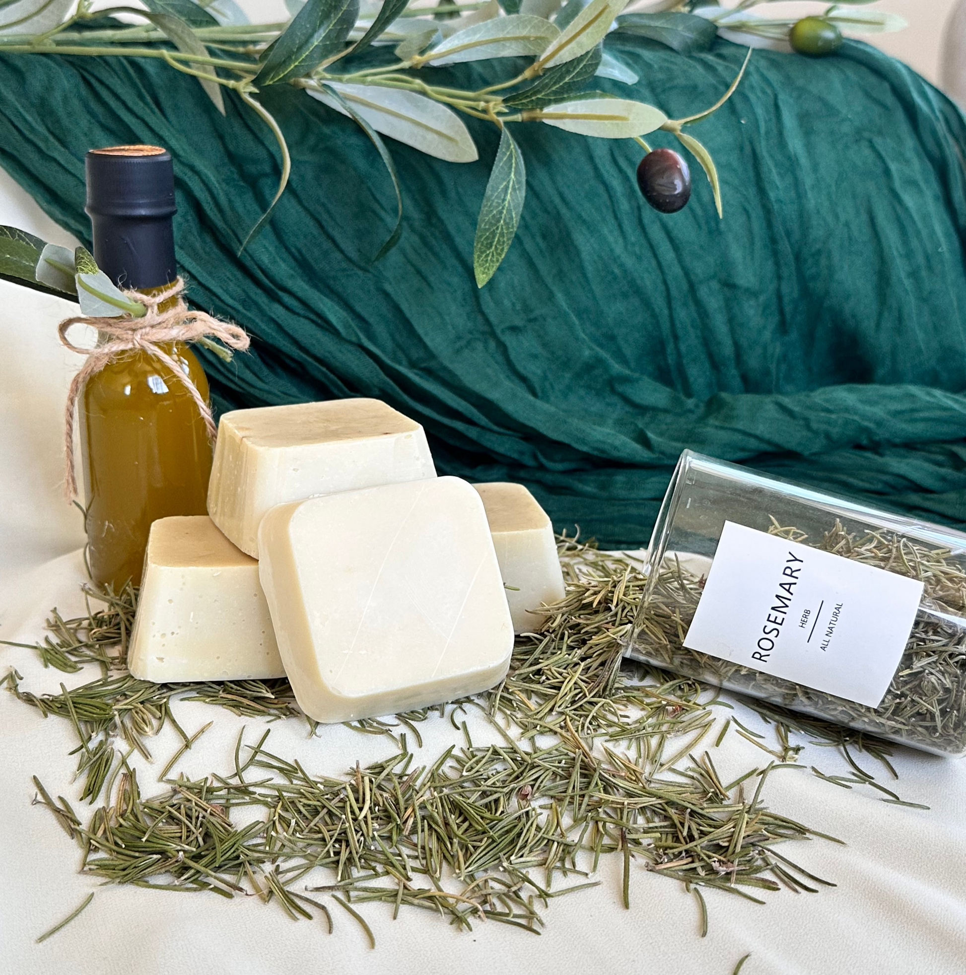 Herbal soap bars and a bottle of oil on a white surface with rosemary leaves.